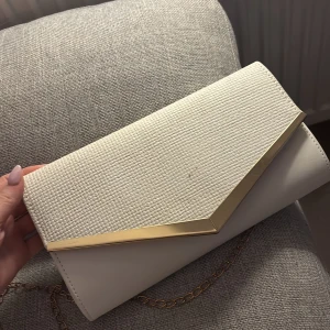 Vit kuvertväska med gulddetaljer - Elegant vit kuvertväska med en stilren guldkant och kedja. Väskan har en praktisk magnetknäppning och en inre ficka för småsaker. Perfekt för att ge en touch av lyx till din outfit. Använd ca 2 gånger på bal. Möjligtvis lite smutsig på utsidan men inget fel på den. Man kan ha de. Bara i handen eller ta ut bandet och sätta den runt axeln. Köpt på SHEIN för 129, mitt pris 60kr. 