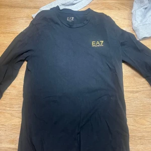 Svart långärmad tröja från EA7 - Säljer en svart långärmad tröja från EA7 Emporio Armani. Tröjan har en enkel design med en liten guldfärgad logotyp på bröstet. Perfekt för en stilren och avslappnad look. Den har en liten skada på armen 