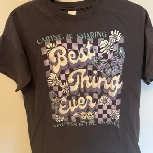 Brun t-shirt med tryck från Lindex - Säljer en brun t-shirt från Lindex med ett färgglatt tryck av texten 'Best Thing Ever' omgiven av blommor och fjärilar. T-shirten har korta ärmar och en rund halsringning. Perfekt för en avslappnad stil!