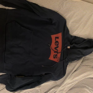 Mörkblå hoodie från Levi's - Säljer en svart hoodie från Levi's med röd logga på framsidan. Tröjan har en klassisk känguruficka och justerbar huva med dragsko. Perfekt för en avslappnad stil. Fint skick. 