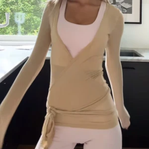 Exercere  - Säljer en stilren beige omlottopp med långa ärmar och knytband i midjan. Perfekt för en elegant och avslappnad look. Storlek M/L men kan passa mindre då man kan knyta själv.