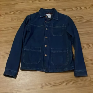 Blå jeansjacka från Jack & Jones - Säljer en snygg blå jeansjacka från Jack & Jones. Jackan har långa ärmar och knappar framtill. Insidan är fodrad med ett rutig mönster i rött, vitt och svart. Perfekt för en casual look!