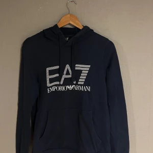 Mörkblå hoodie från Emporio Armani - Säljer en stilren mörkblå hoodie från Emporio Armani med EA7-logga på framsidan. Tröjan har en klassisk design med huva och känguruficka. Perfekt för en avslappnad look.