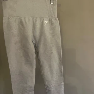 Grå leggings från Gymshark - Säljer ett par grå leggings från Gymshark med hög midja och ribbad detalj upptill. Perfekta för träning med sin stretchiga och bekväma passform. Diskret logga på framsidan. Passar bra för både gym och yoga.