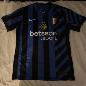 Inter Milan 24/25 hemmatröja  - Säljer en blå och svart randig fotbollströja från Nike med korta ärmar. Tröjan har klubbmärke och sponsorloggor på framsidan samt en Dri-FIT etikett. Perfekt för fotbollsfans som vill visa sitt stöd.