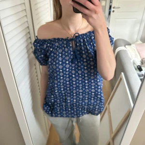 Blå offshoulder blus från Ellos - Säljer en söt blå offshoulder blus från Ellos med vitt mönster. Blusen har korta ärmar och en knytning framtill. Perfekt för en avslappnad stil.