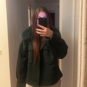 Meotinejacka - Snygg mörkgrön jacka med en stor, fluffig pälskrage. Jackan har knappar framtill och två stora fickor på bröstet. Perfekt för kyligare dagar när du vill hålla stilen. Jackan är från Meotine