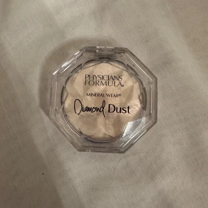 Physicians Formula Mineral Wear Diamond Dust - Säljer en Physicians Formula Mineral Wear Diamond Dust highlighter. Den har en skimrande finish som ger en perfekt glow. Produkten kommer i en transparent, åttakantig förpackning och har en vacker mönstrad yta.
