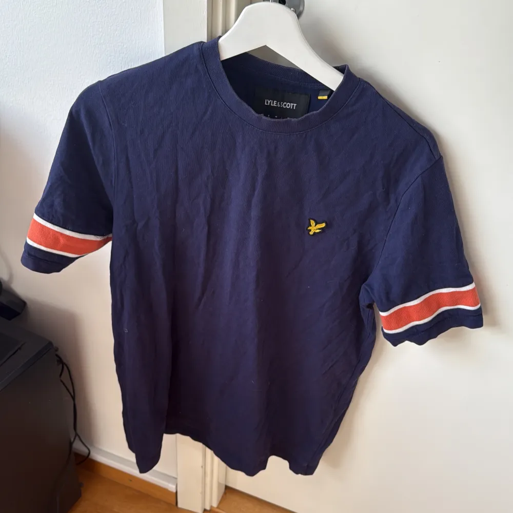 Säljer en mörkblå t-shirt från Lyle & Scott med korta ärmar och ränder i orange och vit på ärmarna. Den har en liten gul logga på bröstet. Perfekt för en avslappnad stil.. T-paidat.