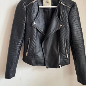 Svart skinnjacka från Vero Moda - Snygg svart skinnjacka från Vero Moda med coola dragkedjedetaljer och quiltade partier på axlarna. Jackan har en klassisk bikerstil med asymmetrisk dragkedja och långa ärmar. Perfekt för att ge din outfit en edgy touch.