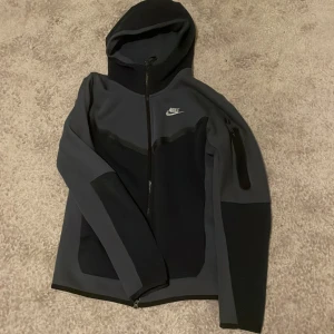 Grå och svart nike tech  - Snygg grå och svart jacka från Nike med huva och dragkedja. Perfekt för träning eller vardagsbruk. Niketechen har en ficka med dragkedja på ärmen och Nike-logga på bröstet. Säljer för att den är för stor på mig. Tveka inte om du har frågor eller undrar något. Skriv direkt.