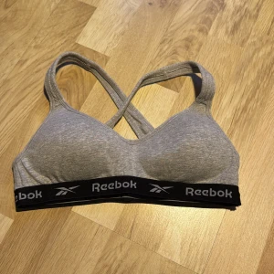 Grå sporttopp från Reebok - Säljer en grå bh från Reebok med korsade axelband och elastiskt band med logga. Perfekt för träning och bekväm passform.
