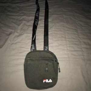 Svart axelväska från Fila - Säljer en stilren svart axelväska från Fila med justerbar rem och dragkedja. Perfekt för att ha mobil plånbok och annat. 