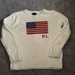 Vit stickad tröja med amerikansk flagga - Säljer en vit stickad tröja från Ralph Lauren med en stor amerikansk flagga på framsidan och initialerna 'RL'. Tröjan har långa ärmar och ribbade kanter vid ärmslut och nederkant. Perfekt för en stilren och avslappnad look.