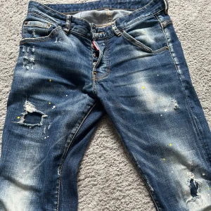 Blå jeans med slitningar - Snygga blå jeans med slitningar och färgstänk för en cool look. De har en klassisk femficksdesign och en knappgylf. Perfekta för en avslappnad stil.