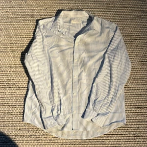 Randig skjorta från Pier One - Snygg och klassisk långärmad skjorta från Pier One i ljusblå och vit randigt mönster. Perfekt för en stilren look. Skjortan har knappar framtill och en bekväm passform. Den är bara använd en gång.