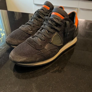 Svarta sneakers med orange detaljer - Snygga svarta sneakers från Philippe Model med orange detaljer och snörning. Skorna har en kombination av mocka och syntetmaterial, vilket ger dem en stilren look. Perfekta för en casual stil med en touch av färg.