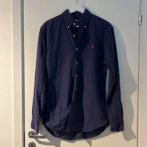 Mörkblå skjorta från Ralph Lauren - Snygg mörkblå skjorta från Ralph Lauren med klassisk krage och knappar framtill. Den har en broderad röd logga på bröstet och långa ärmar. Perfekt för en stilren look.
