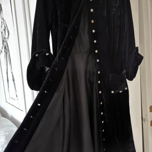 Svart kappa med knappar - Elegant svart kappa i svart plysch med långa ärmar och knappar hela vägen ner. Tillverkad i en stilren design. Perfekt för att ge en sofistikerad touch till din outfit.