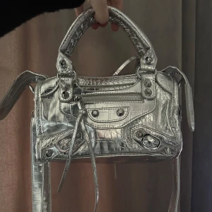 Balenciaga liknande väska - Snygg silverfärgad axelväska med krokodilmönster och dragkedja. Väskan har flera detaljer som knappar och snörning, vilket ger den en unik look. Perfekt för att ge din outfit en extra glans. Dragkedjan kan vara lite seg och dra i men den fungerar som den ska. Bilderna visar skicket