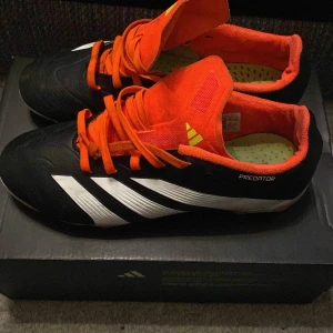 Adidas Predator fotbollsskor - Snygga Adidas Predator fotbollsskor i svart med vita detaljer och orange insida. Skorna har snörning och är designade för optimal prestanda på planen. Perfekta för fotbollsspelare som vill ha stil och funktionalitet.