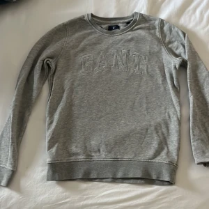 Grå sweatshirt från GANT - Säljer en stilren grå sweatshirt från GANT med broderad logga på bröstet. Tröjan har långa ärmar och rund halsringning, perfekt för en avslappnad look.