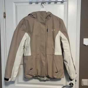 Beige och vit jacka från Oakley - Säljer en snygg beige och vit skidjacka från Oakley. Jackan har en huva och dragkedja framtill. Endast använd fåtal gånger. Normal i storleken, för mig som normalt har storlek s-m är den lite oversize. Nypris 2200kr