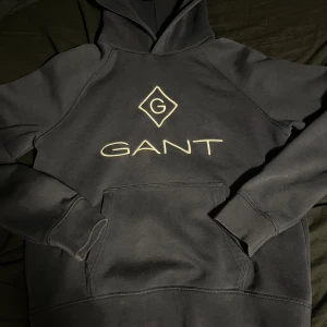 Svart hoodie från GANT - Säljer en stilren svart hoodie från GANT med broderad logga på bröstet. Tröjan har en klassisk känguruficka och långa ärmar. Perfekt för en avslappnad stil.
