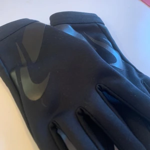 Svarta Nike handskar  - Snygga svarta handskar från Nike med deras ikoniska logotyp i en diskret design. Handskarna har en greppvänlig yta på insidan och är fodrade för extra värme. Det står att det är i storlek M men det passar bättre för S och XS. I mycket bra skick och är utan några skador eller skavanker. Bilder på logo, skötselråd och handskarnas material är inkluderade. Perfekta för kyliga dagar när du vill hålla stilen. Återkom gärna om det finns några funderingar eller om fler bilder begärs ☺️
