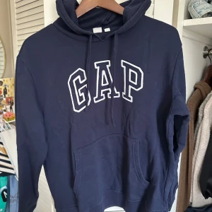 Mörkblå hoodie från GAP - Säljer en snygg mörkblå hoodie från GAP med stor logga på framsidan. Tröjan har en klassisk känguruficka och justerbar huva med dragsko. Perfekt för en avslappnad stil. 🧢