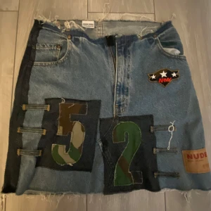 Jeanskjol med armédetaljer från Nude - Unik jeanskjol från Nude med coola armédetaljer. Kjolen har en patch med siffrorna 52 i camouflage och en armélogga med stjärnor. Den har en dragkedja framtill och råa kanter för en edgy look. Perfekt för dig som vill sticka ut! Strl 29