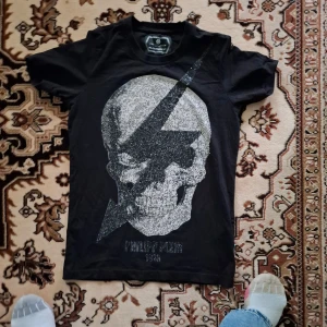 Svart t-shirt med dödskallemotiv från Philipp Plein - Snygg svart t-shirt från Philipp Plein med ett stort glittrigt dödskallemotiv och blixt på framsidan. Perfekt för dig som vill sticka ut med en cool och edgy stil. T-shirten har korta ärmar och en klassisk passform.