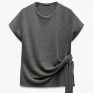 Grå stickad topp med knyt från Zara - Mörkgrå stickad topp från Zara med korta ärmar och rund hals. Toppen har en snygg knytdetalj i sidan som ger en cool och avslappnad look. Perfekt att styla med jeans eller kjol för en trendig vibe.