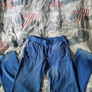 Juicy Couture blå mjukisbyxor - Blå mjukisbyxor från Juicy Couture i supermjuk material med broderad JC-logga på benet. Byxorna har snörning i midjan, stora fickor fram och en bakficka med tryckknappar. Perfekta för chill eller streetwear.
