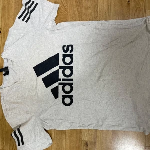 Grå Adidas t-shirt med svart logga - Snygg ljusgrå t-shirt från Adidas med stor svart logga framtill och klassiska svarta ränder på ärmarna. T-shirten har rund hals och är tillverkad i mjuk bomull, perfekt för en sportig och avslappnad stil.