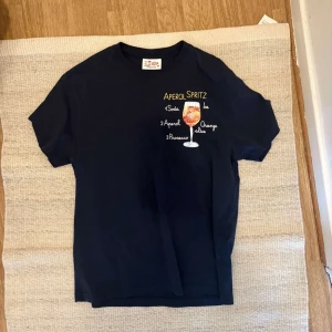 Saint barth t shirt - Mörblå t-shirt från ST barth med färgglad Aperol Spritz-tryck och recept på bröstet. Klassisk passform, rund hals och korta ärmar. Perfekt för dig som gillar coola och roliga prints. Gjord i mjuk bomull för skön känsla. Aldrig använd. Nyskick!