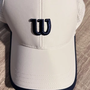 Vit keps från Wilson S/M - Snygg vit keps från Wilson med svart broderad W-logga framtill. Klassisk böjd skärm och ventilerande perforeringar på sidorna. Perfekt för sportiga dagar eller när du vill ha en clean look. Modellen heter Structured Classic Cap i färgen Sanddrift.
