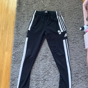 Svarta Adidas träningsbyxor med vita ränder - Säljer ett par svarta Adidas träningsbyxor med klassiska vita ränder längs sidorna och nummer 5 tryckt på låret. Byxorna har dragsko i midjan, dragkedja vid benslut och är gjorda i ett mjukt och sportigt material. Perfekta för träning eller chill. Pris kan diskuteras.