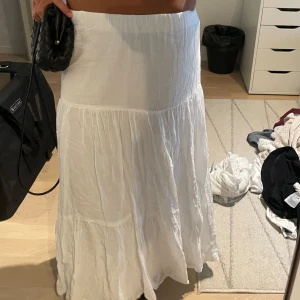 Vit långkjol i bomull med volang - Säljer en superfin vit långkjol i luftig bomull. Kjolen har flera volanglager som ger en flowy och avslappnad vibe. Perfekt för varma dagar när du vill ha något bekvämt men ändå snyggt. Enkel att matcha med crop tops eller stickade tröjor.