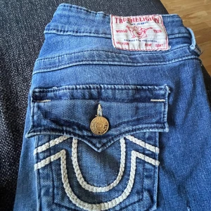 True Religion Joey Low Rise Flare Jeans - Säljer ett par blå True Religion Joey jeans med låg midja och flare-ben. Köpta för 1800kr. Skulle kunna tänka mig att gå ner i pris förmodligen.  Ganska använda. Byxorna sprack vid byx grenen som man ser på sista bilderna men jag har lämnat in dom så dom blev fixade men man ser att dom är lagade ifall man skulle kolla underifrån. Dom är även lite slitna längst ner vid på ena benet men det märker man inte
