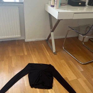 Svart croppad kofta från BikBok - Svart croppad kofta från BikBok med långa ärmar och volangkant vid ärmslut och nederkant. Tröjan har knappar framtill och är i ett mjukt, stretchigt material som känns skönt mot huden. Perfekt att styla med jeans eller kjol.