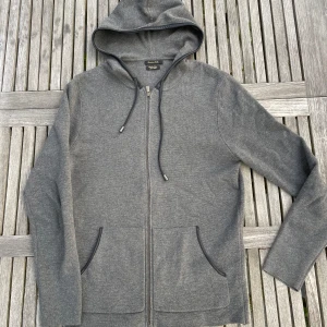 Grå hoodie från Massimo Dutti - Snygg grå hoodie från Massimo Dutti i bomull med dragkedja och huva. Tröjan har svarta detaljer runt huvan och snörning, samt två fickor framtill. Perfekt för en chill och stilren look.