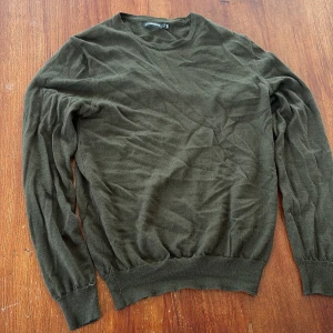 Olivgrön stickad tröja J.Lindeberg - Snygg olivgrön stickad tröja från J.Lindeberg med rund halsringning och diskret logga på bröstet. Tröjan har långa ärmar och ribbade muddar vid ärmslut och nederkant. Perfekt för lager-på-lager och chill stil.