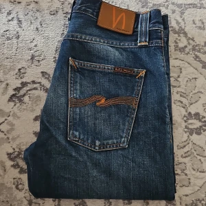 Nudie Jeans Steady Eddie Whistle Blue - Snygga Steady Eddie jeans från Nudie Jeans i tvätten Whistle Blue. Klassisk femficksmodell med orange kontrastsömmar och ikonisk brodyr på bakfickorna. Tillverkade i 100% ekologisk bomull för en schysst känsla och look.