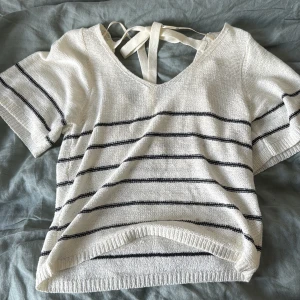 Randig stickad topp med knyt från JDY - Superfin vit stickad topp från JDY med svarta ränder och kort ärm. Snygg v-ringning och knytband i ryggen som ger en extra detalj. Perfekt till jeans eller kjol för en chill look. Mjuk och skön, passar perfekt till vår och sommar.