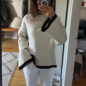 Vit stickad tröja med svarta detaljer - Snygg vit stickad tröja med breda, utsvängda ärmar och svarta kontrastkanter vid ärmslut och nederkant. Tröjan har en avslappnad passform och ribbade detaljer som ger en cool och modern look. Perfekt att styla med jeans eller kjol.