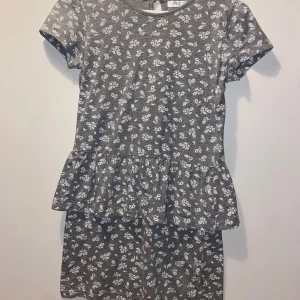 Grå blommig klänning från bpc - Supersöt grå kortklänning från bpc med vitt blommönster, volang i midjan och knapp i nacken. Mjuk och skön bomullskänsla, perfekt för dig som gillar en chill och söt stil.