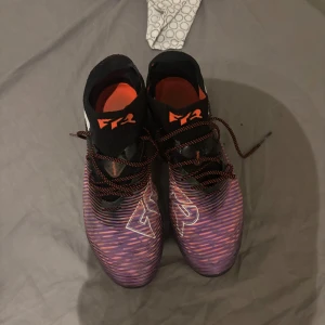 Puma Future Z fotbollsskor svart/lila - Säljer ett par Puma Future Z fotbollsskor i storlek 40.5. Skorna har en cool svart och lila design med orange detaljer och snörning. Ovandelen är i syntet och stickat material för bra passform och känsla på planen. Sulan har dobbar för optimalt grepp.
