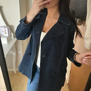 Mörkblå trenchcoat i denim - Säljer en snygg mörkblå skepparkavaj i jeansmaterial med dubbelknäppning och bred krage. Jackan har en loose passform och stora svarta knappar framtill. Perfekt för dig som gillar en chill och stilren look.