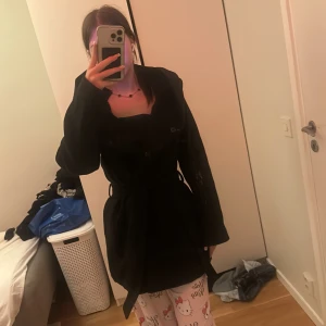 Svart kappa från Amisu med bälte - Svart kappa från Amisu med snyggt bälte i midjan och stora slag. Klassisk och stilren modell med knappar framtill. Perfekt för dig som gillar en enkel men elegant look. Fodrad insida för extra komfort.